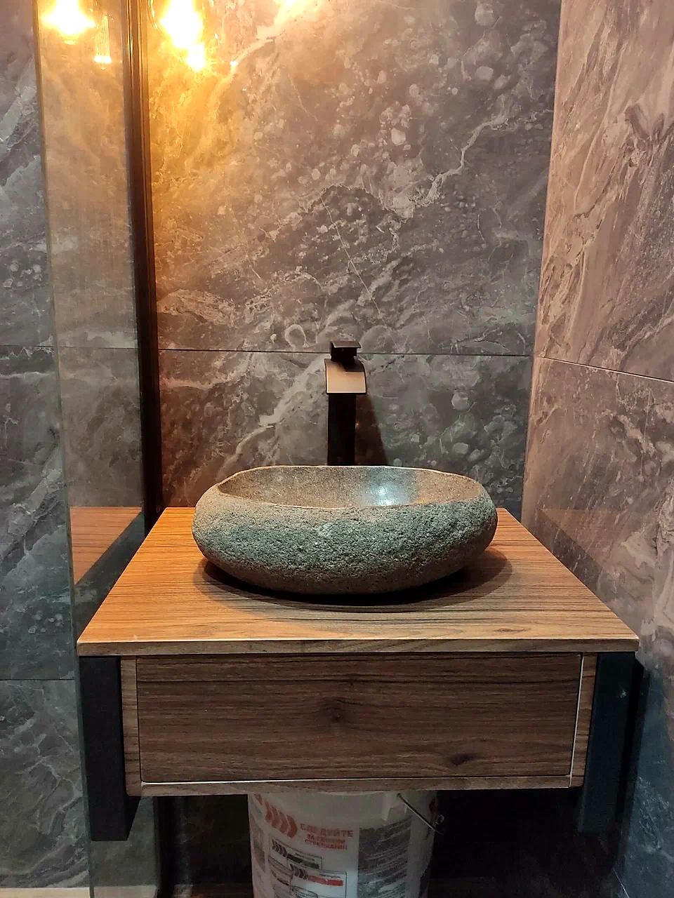 Dark gray stone vessel sink on a natural wood vanity with a large mirror and black brick wall